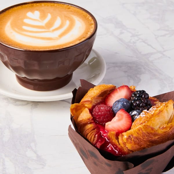 A latte with foam art and a berry pastry in a brown wrapper on a marble surface.