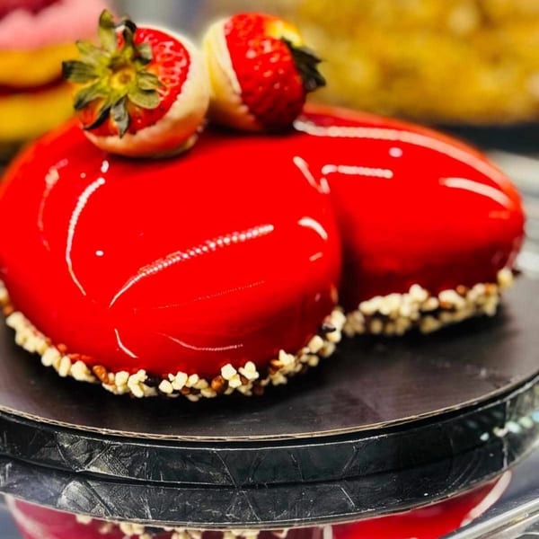 A heart-shaped red glazed cake with white chocolate-dipped strawberries and nut crumble.